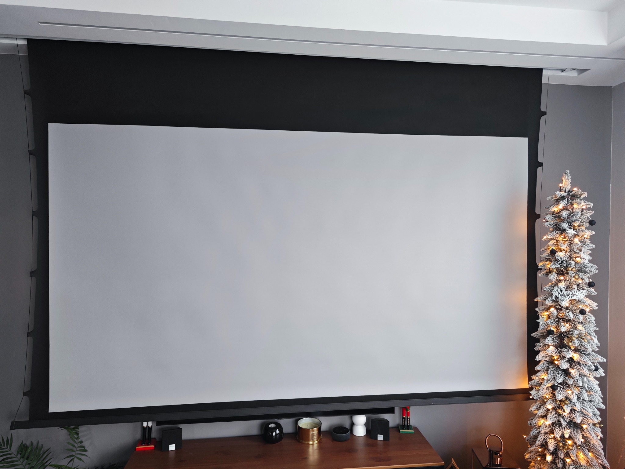 Automatycznie rozwijany ekran projekcyjny w szarej obudowie, zamontowany pod sufitem, z dekoracjami na szafce pod ekranem i choinką z boku, tworzący domowe kino.