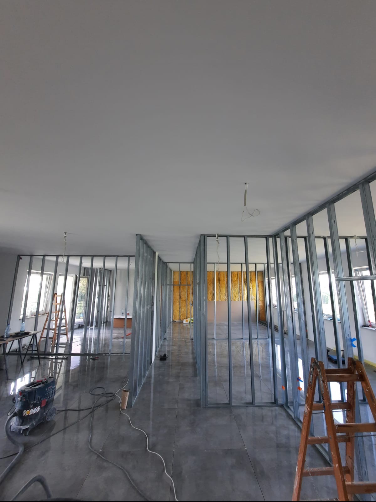 Wnętrze w trakcie remontu: metalowe profile ścian działowych, częściowo wypełnione wełną mineralną, błyszcząca podłoga, drabina i odkurzacz przemysłowy.