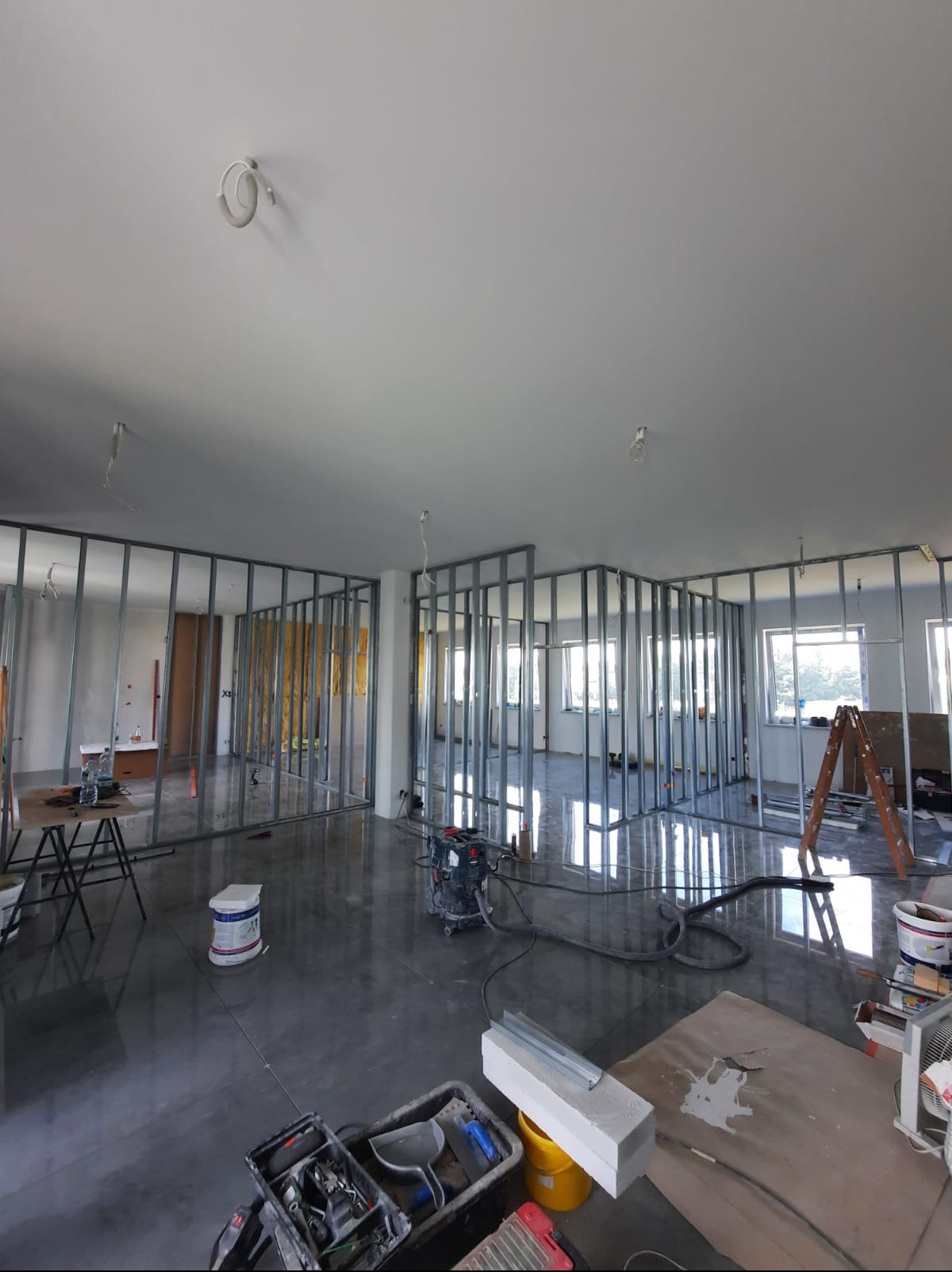Stelaż z profili metalowych tworzący konstrukcję ścian działowych w surowym stanie, widoczny odkurzacz przemysłowy, narzędzia i drabina, posadzka z wylewki samopoziomującej.