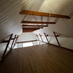 Michał Apon - Wykończone poddasze z drewnianą podłogą, białą boazerią na skosach dachu i widocznymi drewnianymi belkami konstrukcyjnymi.
