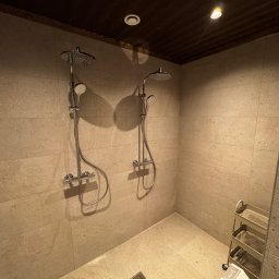 Michał Apon - Dwa zestawy prysznicowe z deszczownicą i słuchawką, zamontowane na ścianie z płytek w odcieniach beżu w łazience w Rogoźnie, z drewnianym sufitem i metalowym wózkiem na kosmetyki w rogu.