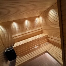 Michał Apon - Wnętrze sauny wykończone jasnym drewnem, z trzypoziomowymi ławkami i piecem Harvia w rogu, oświetlone punktowymi lampami sufitowymi.