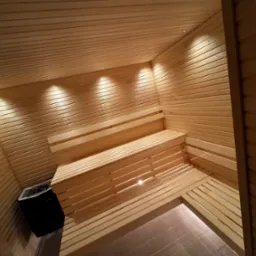 Wnętrze sauny wykończone jasnym drewnem, z trzypoziomowymi ławkami i piecem Harvia w rogu, oświetlone punktowymi lampami sufitowymi.