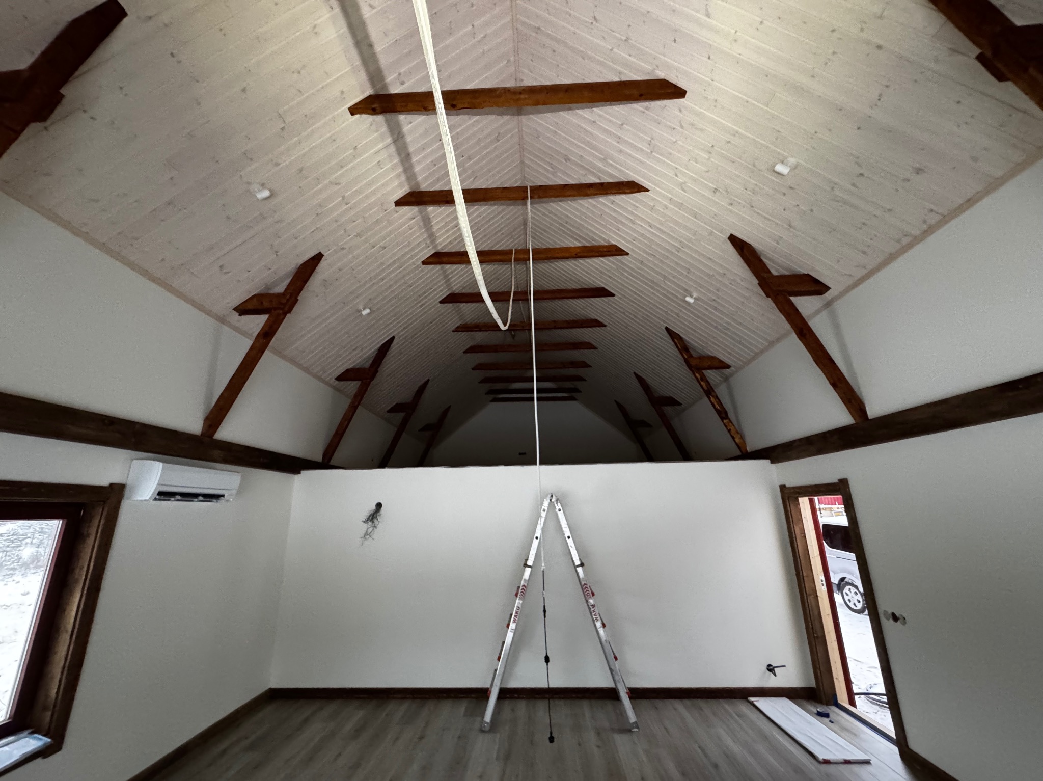 Wnętrze poddasza z drewnianą konstrukcją dachu, białe ściany, klimatyzator, aluminiowa drabina rozstawiona na podłodze, widok na zewnątrz przez otwarte drzwi.
