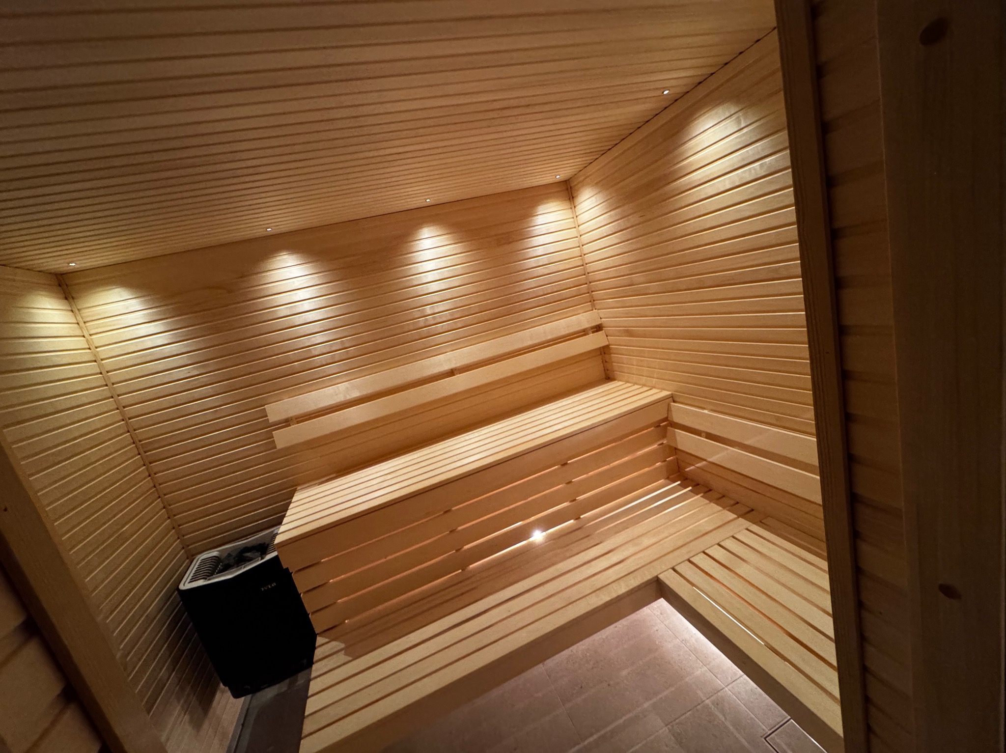 Wnętrze sauny wykończone jasnym drewnem, z trzypoziomowymi ławkami i piecem Harvia w rogu, oświetlone punktowymi lampami sufitowymi.