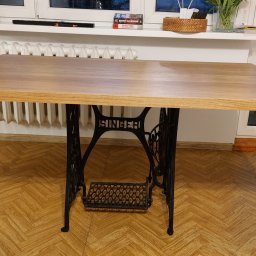 Piotr Andrzejewski - Blat drewniany na metalowej podstawie starej maszyny do szycia Singer. Jasne drewno, widoczne słoje. W tle okno i biały grzejnik.