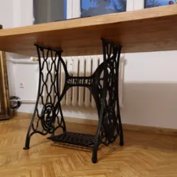 Stół z blatem z jasnego drewna, wsparty na czarnej, ozdobnej podstawie od starej maszyny do szycia Singer. W tle okno i lustro w złotej ramie.