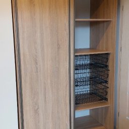 Piotr Andrzejewski - Jasna szafa przesuwna z drewnopodobnymi frontami i metalowymi koszami. Widoczna podłoga z paneli. Minimalistyczny design.