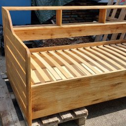 Piotr Andrzejewski - Dębowa, surowa rama łóżka z listewkowym stelażem, ustawiona na palecie. Widoczna faktura drewna i naturalne usłojenie. Tło stanowią materiały budowlane.