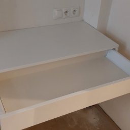 Piotr Andrzejewski - Biała szafka z wysuniętą szufladą, zamontowana pod blatem z gniazdkami elektrycznymi, w surowym wnętrzu. Widoczne niedoskonałości wykończenia ścian.