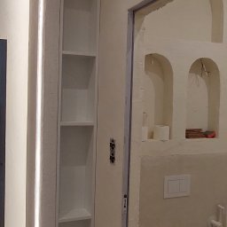 Piotr Andrzejewski - Wąska, wnękowa szafka z oświetleniem LED w łazience w Warszawie. Białe półki, minimalistyczny design, widok na toaletę w tle. Nietypowe wykończenie ścian.