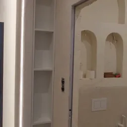 Wąska, wnękowa szafka z oświetleniem LED w łazience w Warszawie. Białe półki, minimalistyczny design, widok na toaletę w tle. Nietypowe wykończenie ścian.