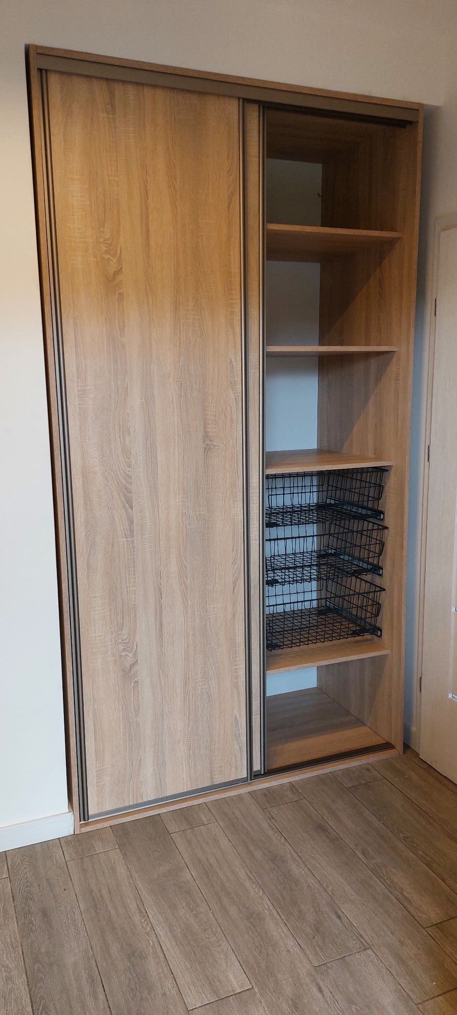 Jasna szafa przesuwna z drewnopodobnymi frontami i metalowymi koszami. Widoczna podłoga z paneli. Minimalistyczny design.