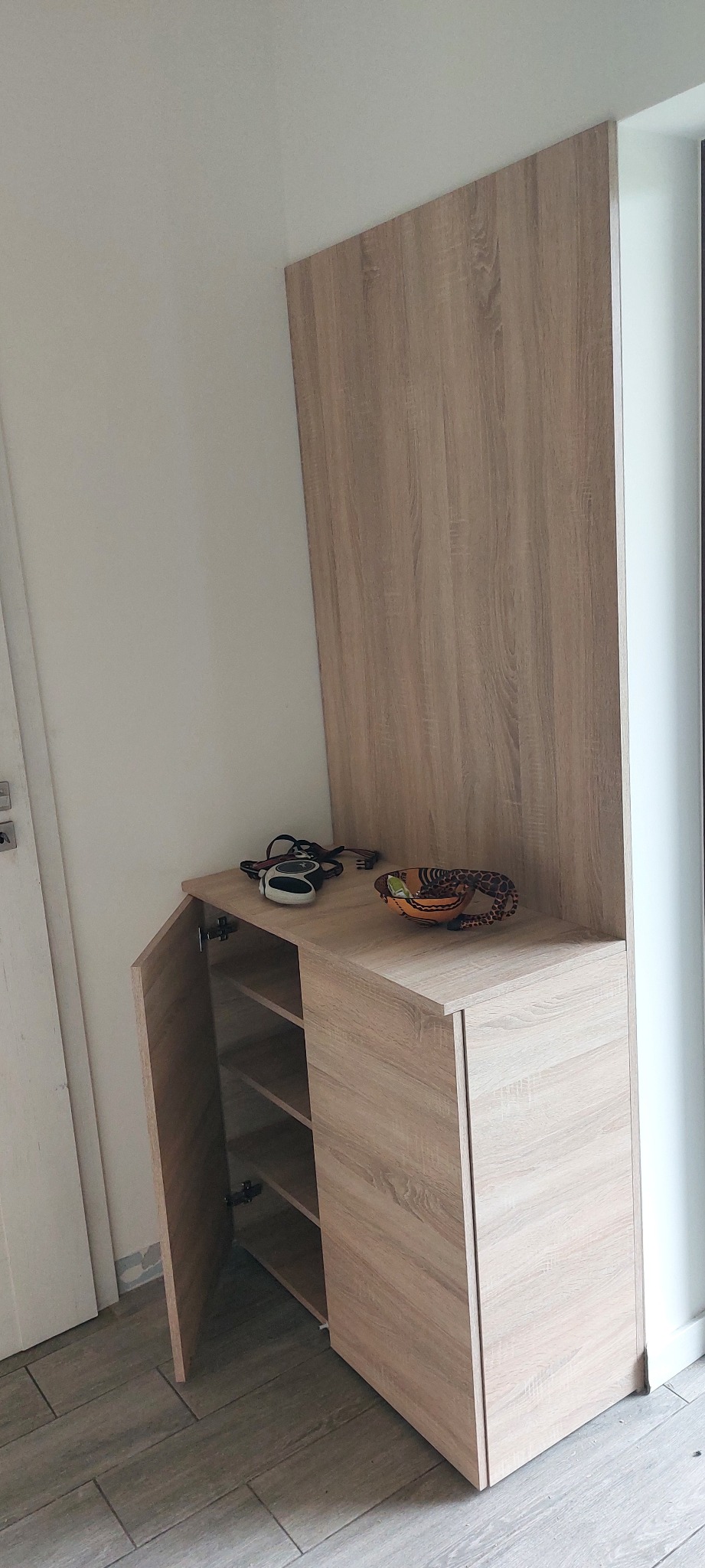 Drewniana szafka na buty z półkami i otwartymi drzwiczkami, z dekoracjami na blacie, w jasnym wnętrzu. Widoczne zawiasy i struktura drewna.