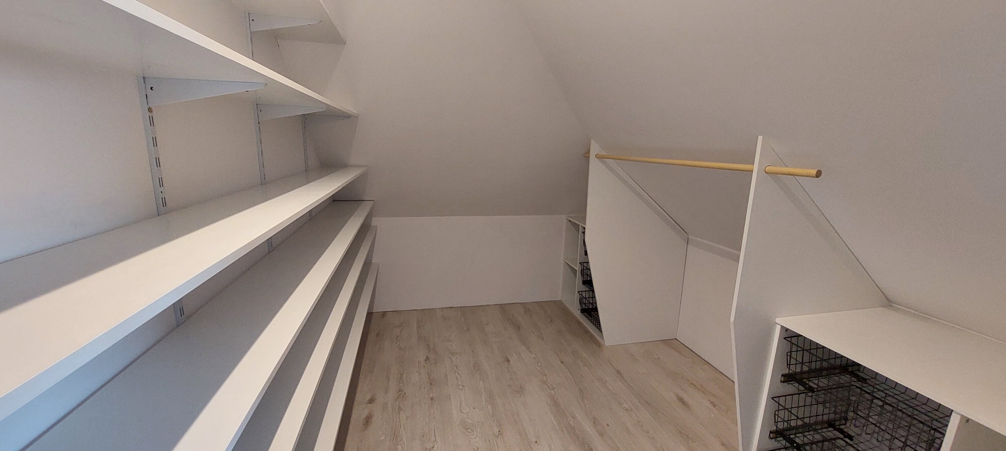 Biała zabudowa pod skosem na poddaszu z półkami i drążkiem na ubrania. Metalowe kosze wkomponowane w zabudowę. Jasna podłoga.