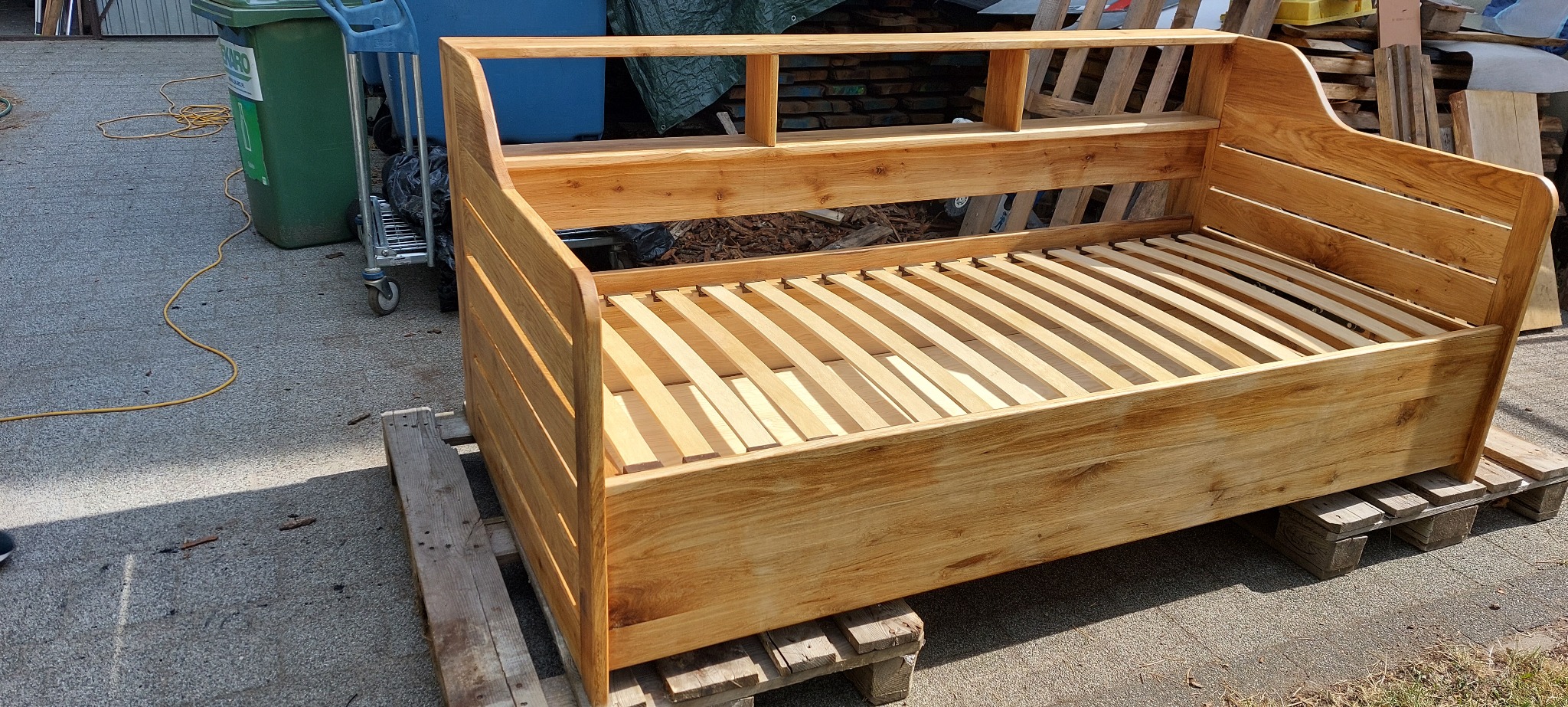 Dębowa, surowa rama łóżka z listewkowym stelażem, ustawiona na palecie. Widoczna faktura drewna i naturalne usłojenie. Tło stanowią materiały budowlane.