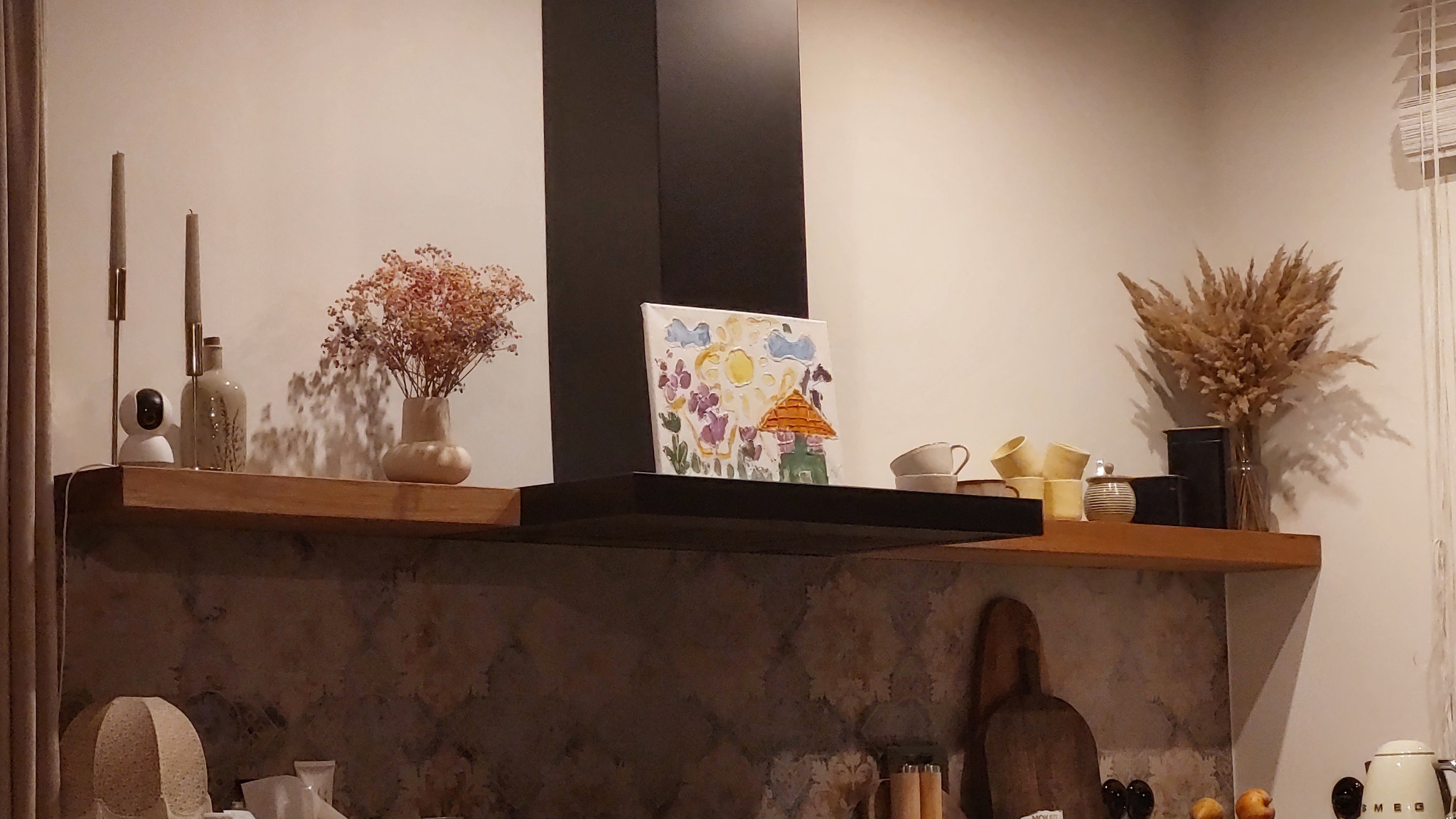Drewniana półka w kuchni z dekoracjami: wazony z suszonymi kwiatami, świece, filiżanki i dziecięcy obrazek. Półka zamontowana pod czarnym okapem.
