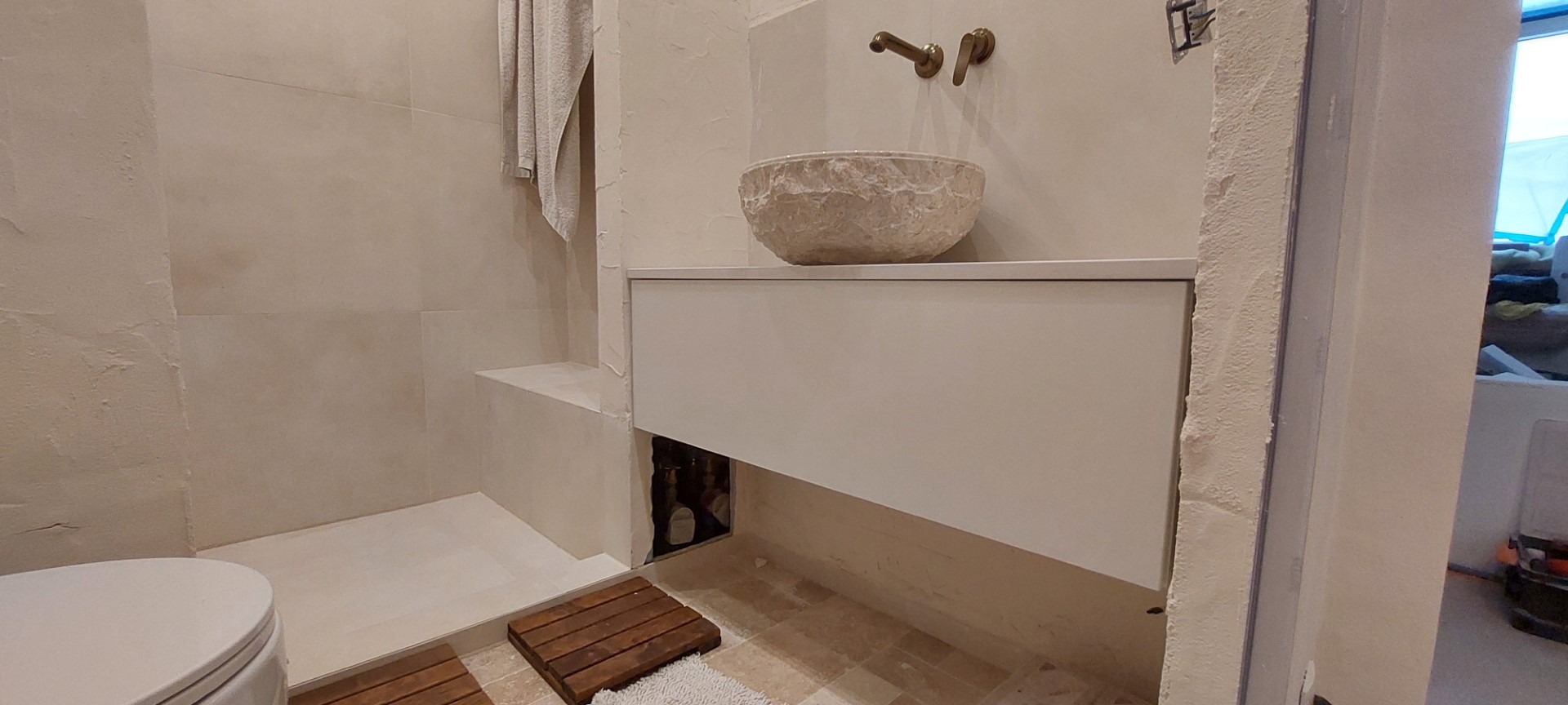 Nowoczesna łazienka w beżach: kamienna umywalka nablatowa, biała szafka pod umywalką i drewniane maty podłogowe. Widoczne fragmenty toalety i niedokończonego wykończenia.