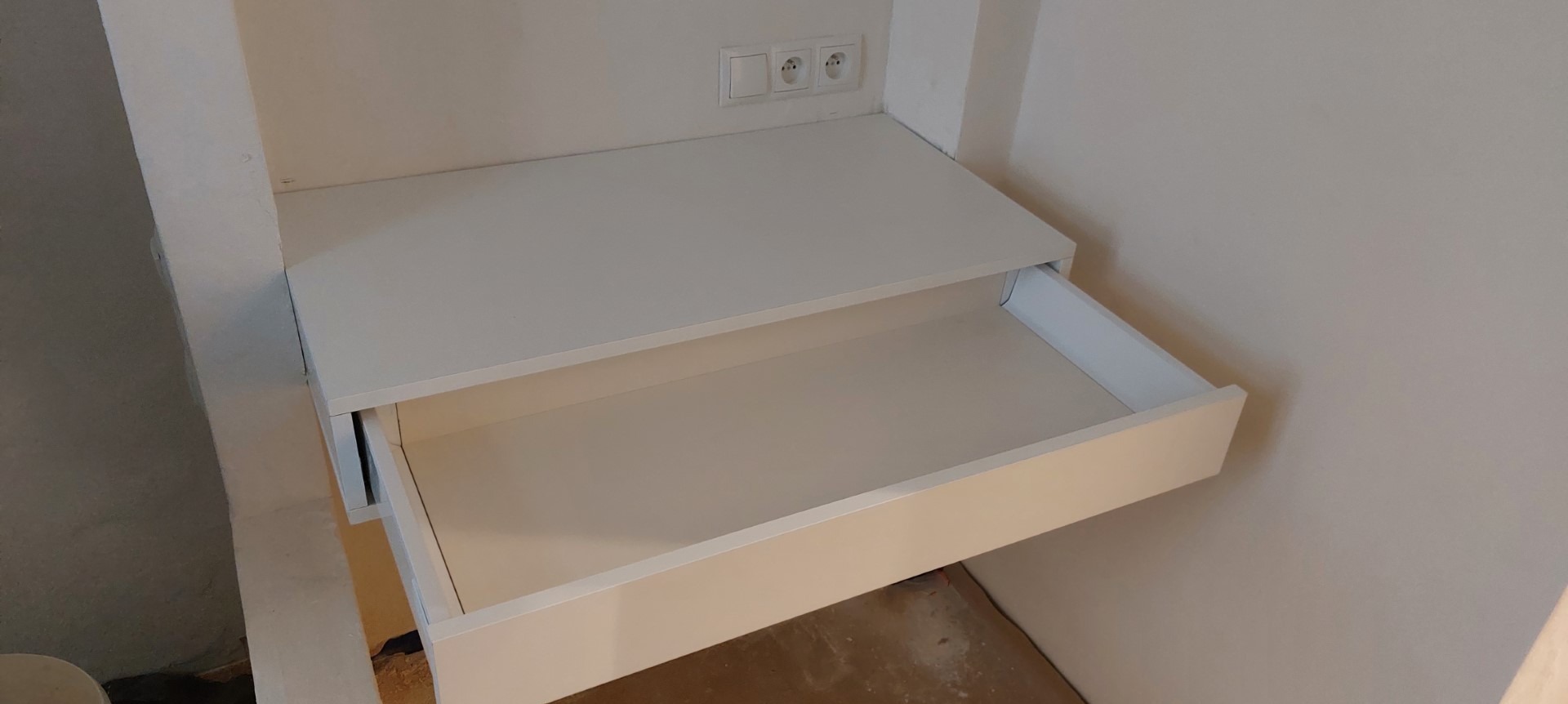 Biała szafka z wysuniętą szufladą, zamontowana pod blatem z gniazdkami elektrycznymi, w surowym wnętrzu. Widoczne niedoskonałości wykończenia ścian.
