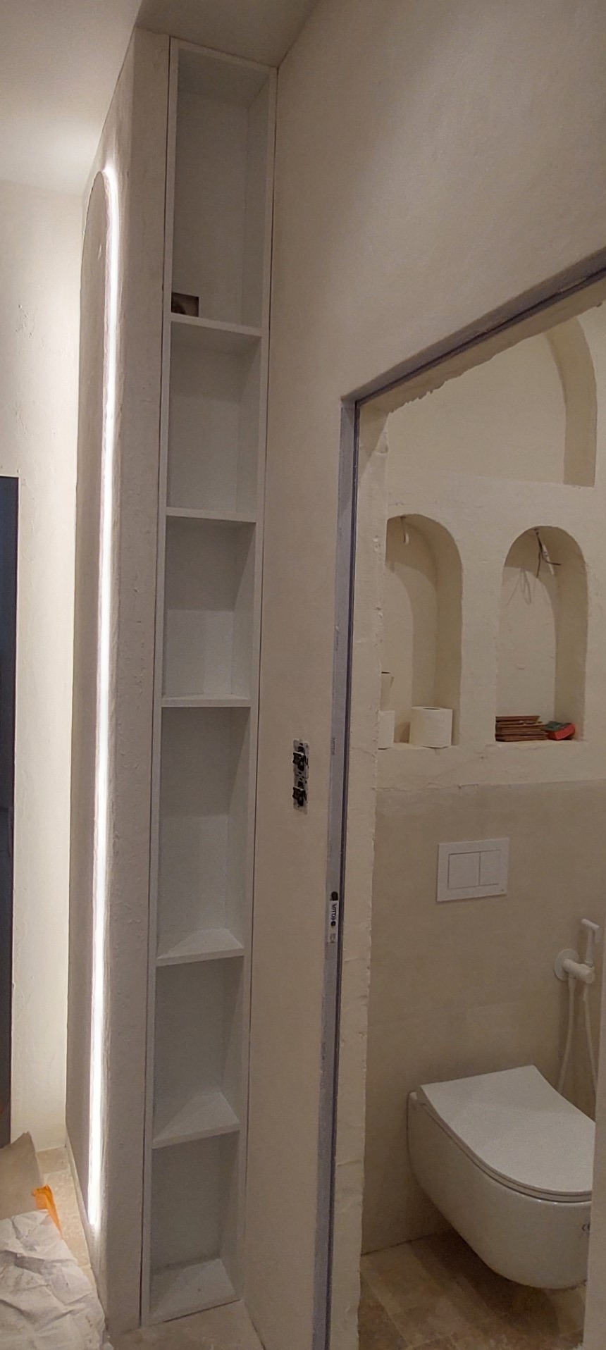 Wąska, wnękowa szafka z oświetleniem LED w łazience w Warszawie. Białe półki, minimalistyczny design, widok na toaletę w tle. Nietypowe wykończenie ścian.