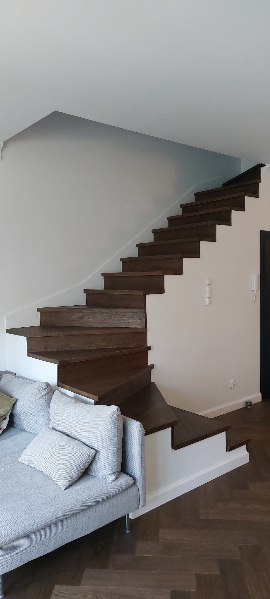 Drewniane schody w nowoczesnym wnętrzu, widoczne z perspektywy salonu. Ciemne stopnie kontrastują z białą ścianą i szarą sofą. Parkiet ułożony w jodełkę.