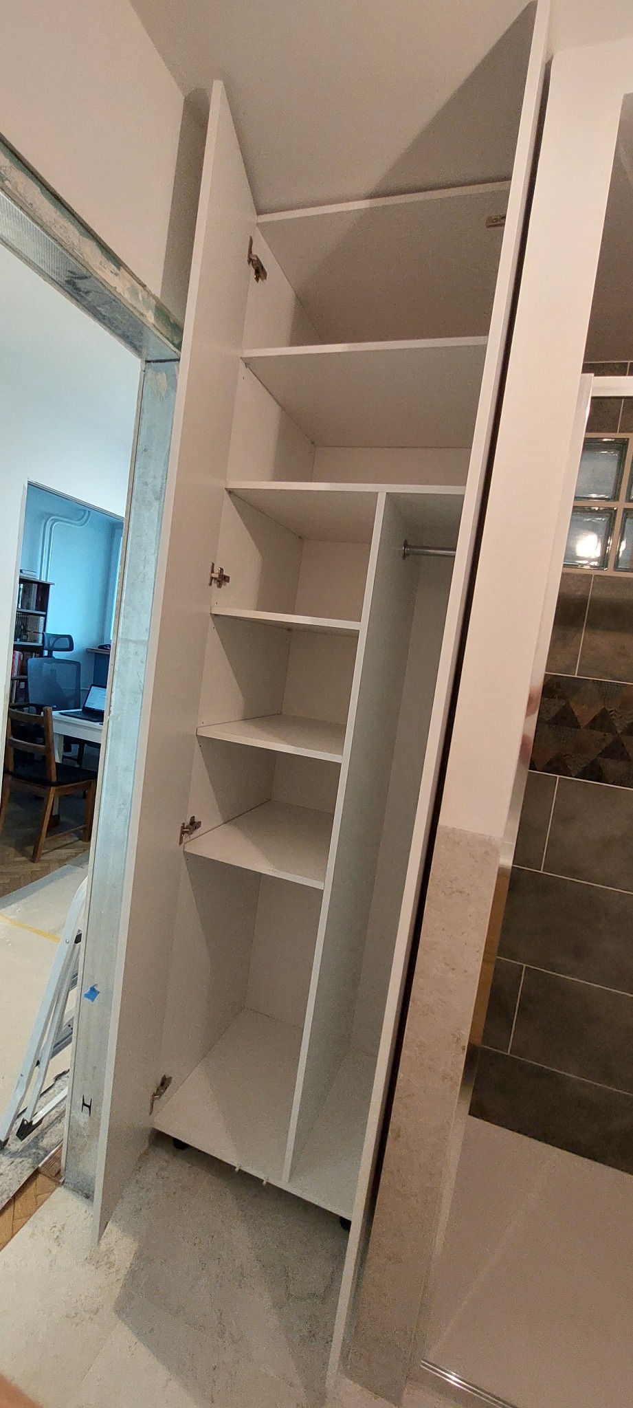 Biała szafa wnękowa z półkami, widoczne zawiasy i drążek na ubrania, wnętrze wykończone, w tle fragment pokoju z biurkiem i krzesłem, oraz fragment łazienki.