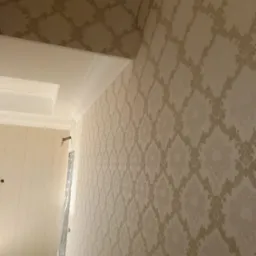 Elegancka ściana w beżowych tapetach z ornamentem, widok z dołu, podkreślony sztukaterią sufitową i listwami przypodłogowymi. W tle ściana w jasnych, gładkich tapetach.