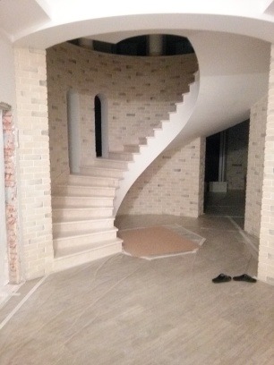 Spiralne schody z ceglanymi ścianami w jasnym wnętrzu. Podłoga zabezpieczona folią malarską, widoczne czarne klapki. Nietypowy kształt schodów.