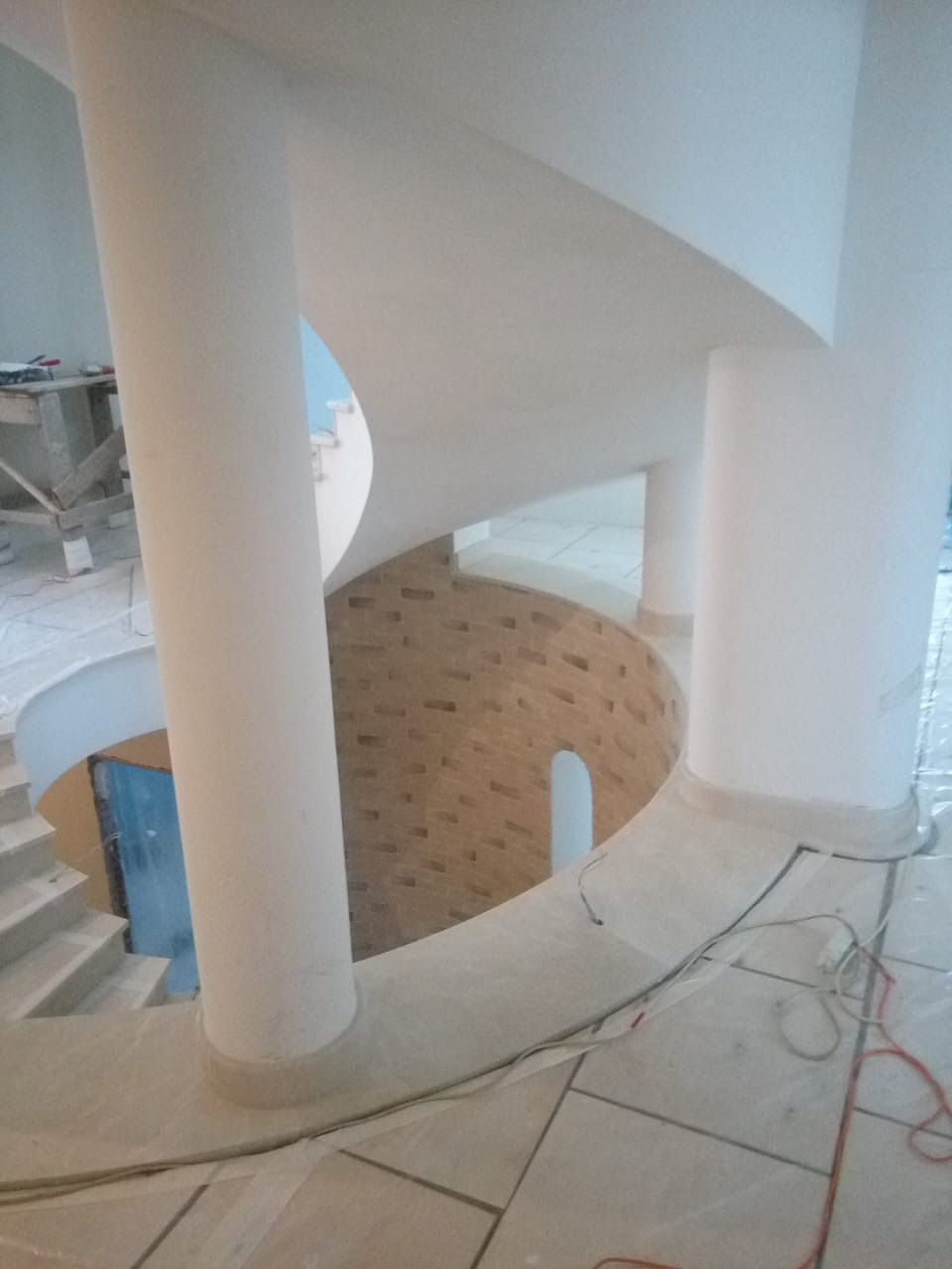 Spiralne schody w trakcie wykańczania, widoczne kolumny i fragment muru z cegły. Prace wykończeniowe wewnątrz budynku.