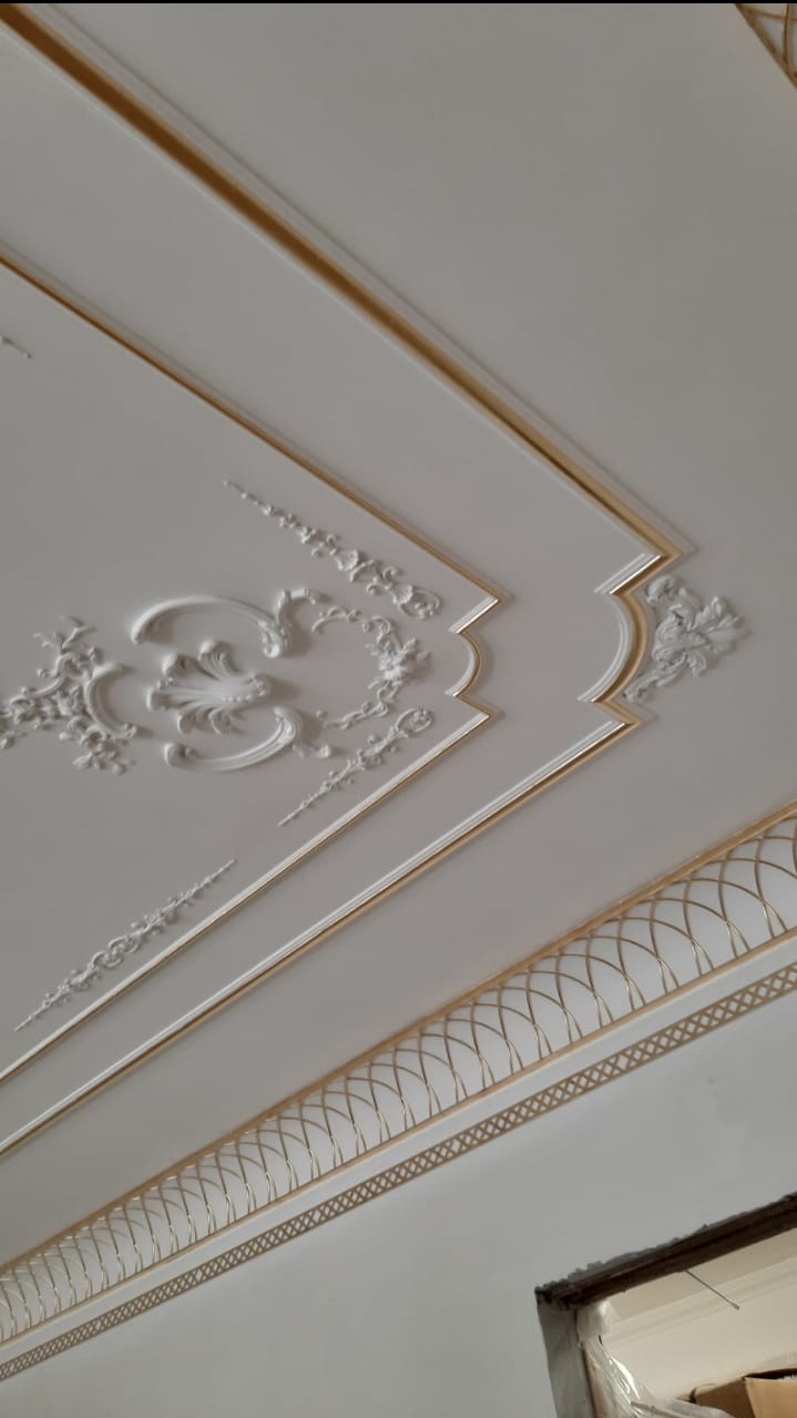 Biały sufit z dekoracyjnymi, gipsowymi zdobieniami i złotymi akcentami, widziany pod kątem. W tle fragment futryny drzwi.