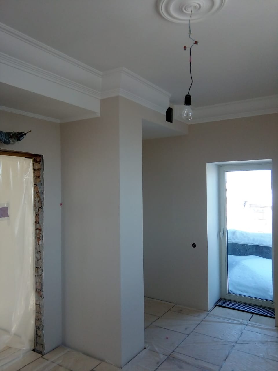 Wykończone ściany z listwami dekoracyjnymi przy suficie w jasnym wnętrzu. Widoczna niedokończona instalacja oświetleniowa i okno z widokiem na śnieg.