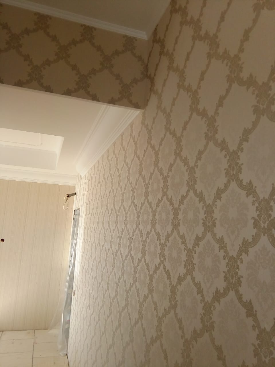 Elegancka ściana w beżowych tapetach z ornamentem, widok z dołu, podkreślony sztukaterią sufitową i listwami przypodłogowymi. W tle ściana w jasnych, gładkich tapetach.