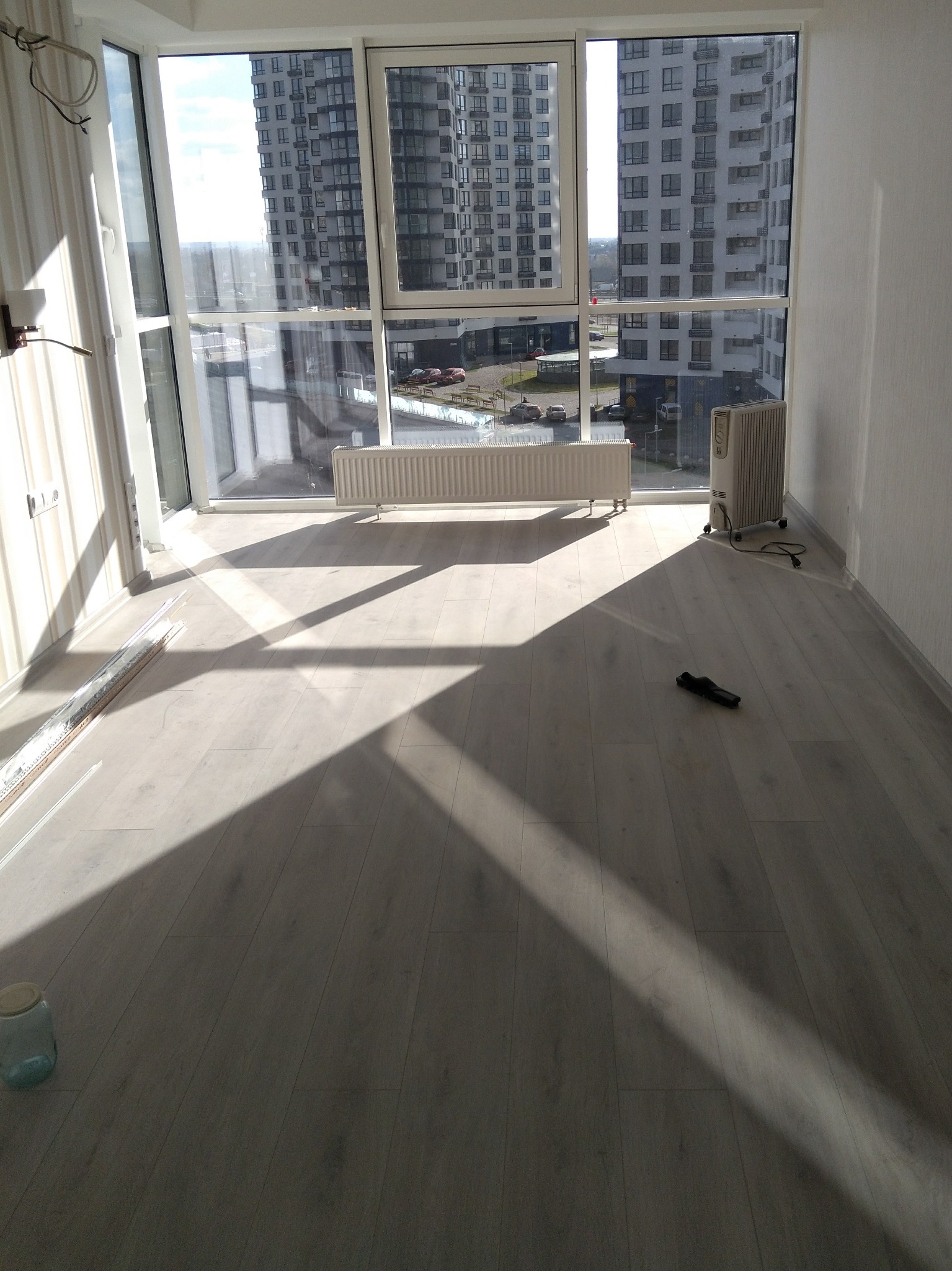 Jasne panele podłogowe w pustym pokoju z dużym oknem i widokiem na budynek. Grzejnik i przenośny grzejnik elektryczny przy ścianie. Promienie słoneczne wpadają do wnętrza.