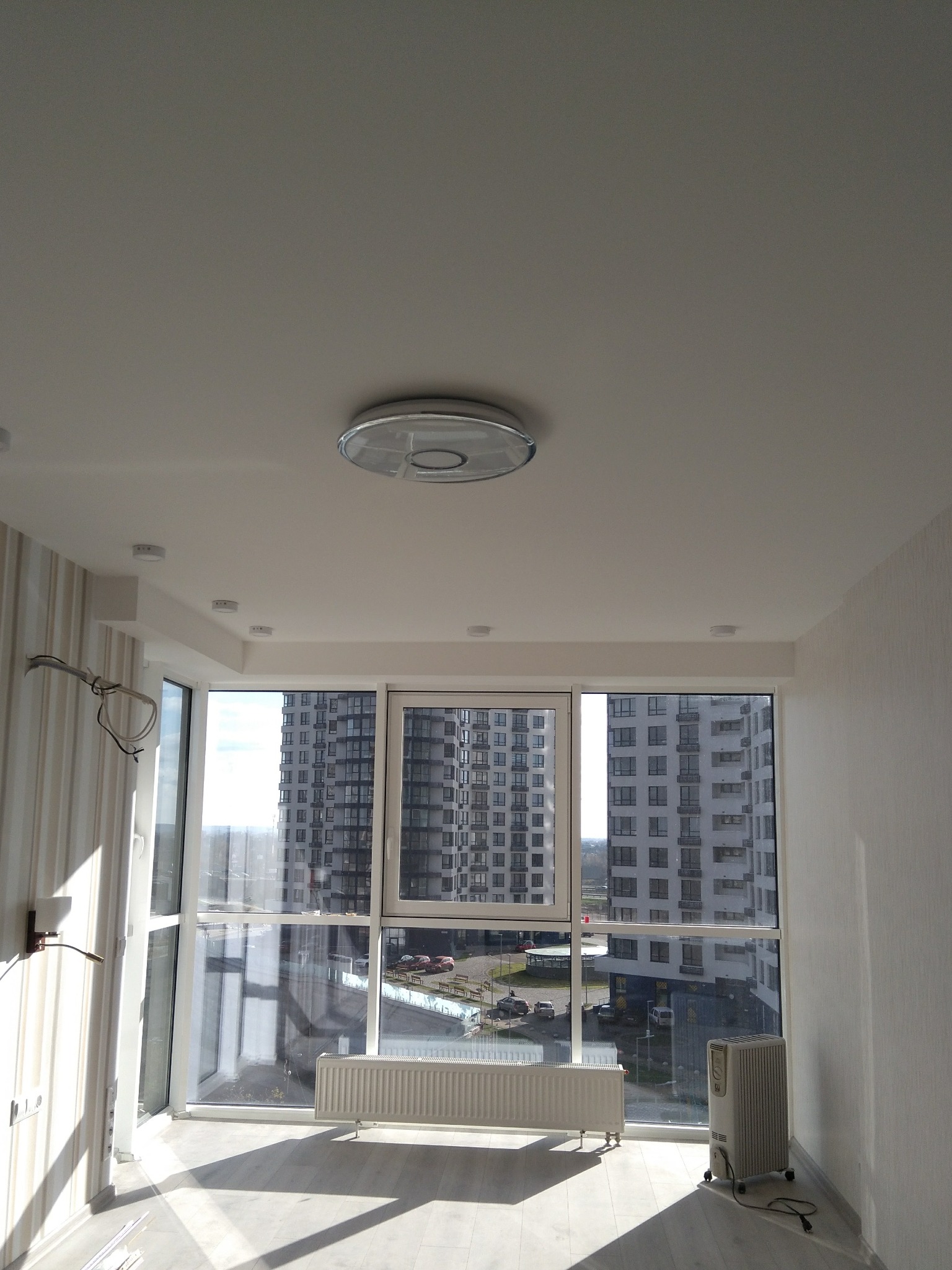 Jasne wnętrze z dużymi oknami i widokiem na budynki. Świeżo wykończone ściany, sufit z oświetleniem punktowym i grzejnik. Minimalistyczny styl, dużo światła dziennego.