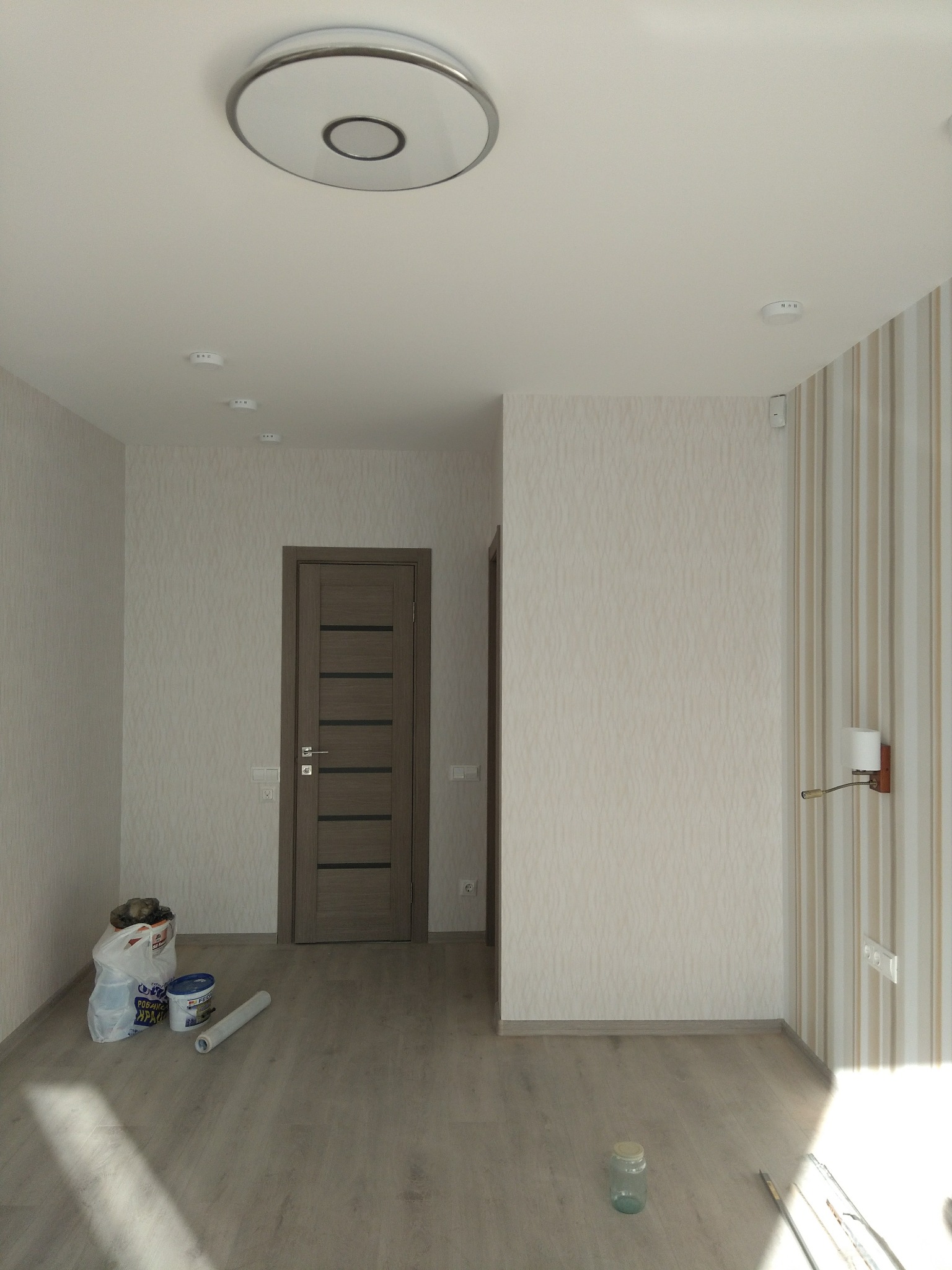 Jasne wnętrze po odświeżeniu: ściany w beżowych tapetach, drewniane drzwi, lampa sufitowa. Na podłodze widoczne akcesoria malarskie i puszka farby.