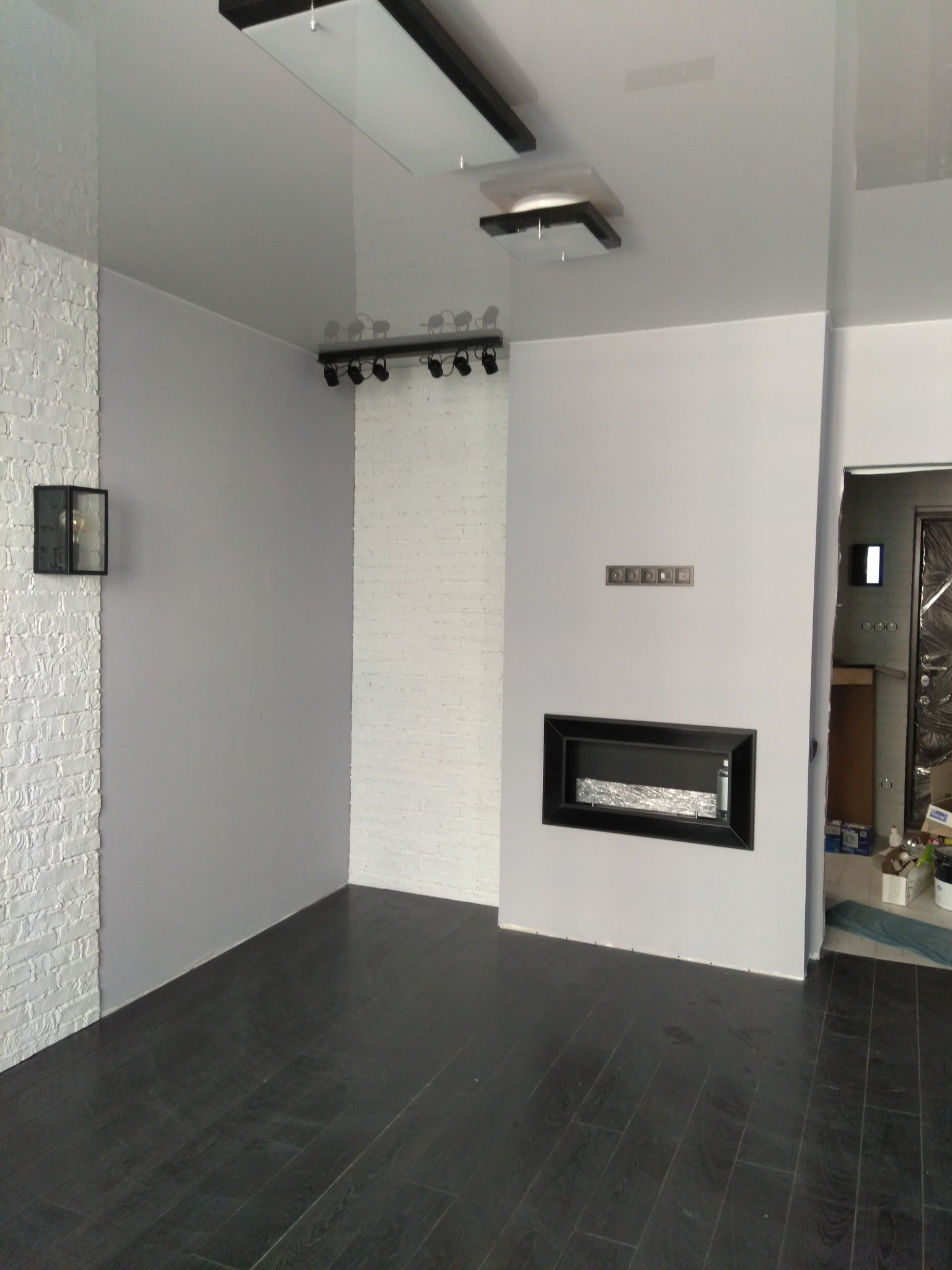 Nowoczesne wnętrze po remoncie: czarna podłoga, biała cegła, szare ściany, minimalistyczny kominek i oświetlenie sufitowe. Widok na wykończony pokój.