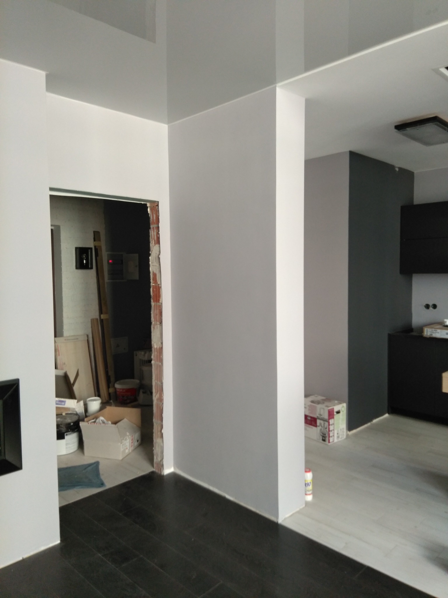 Wnętrze po malowaniu ścian na szaro i biało, widoczne fragmenty kuchni i pokoju w trakcie remontu, ciemna podłoga. Częściowo wykończone pomieszczenie.