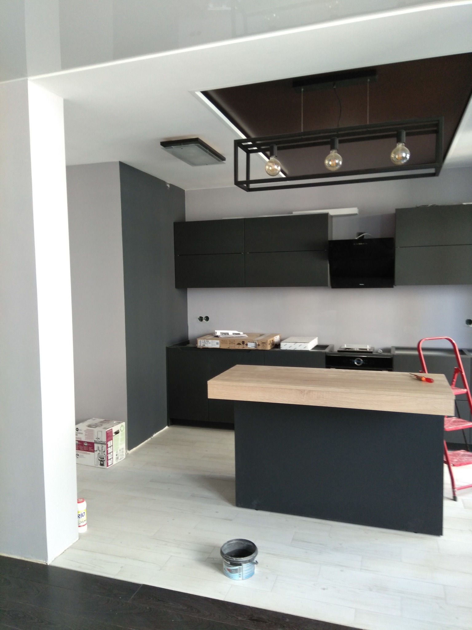 Nowoczesna kuchnia w trakcie wykończenia: ciemne szafki, drewniany blat wyspy, sufit z oświetleniem. Widoczne farby i drabina, co sugeruje etap prac wykończeniowych.