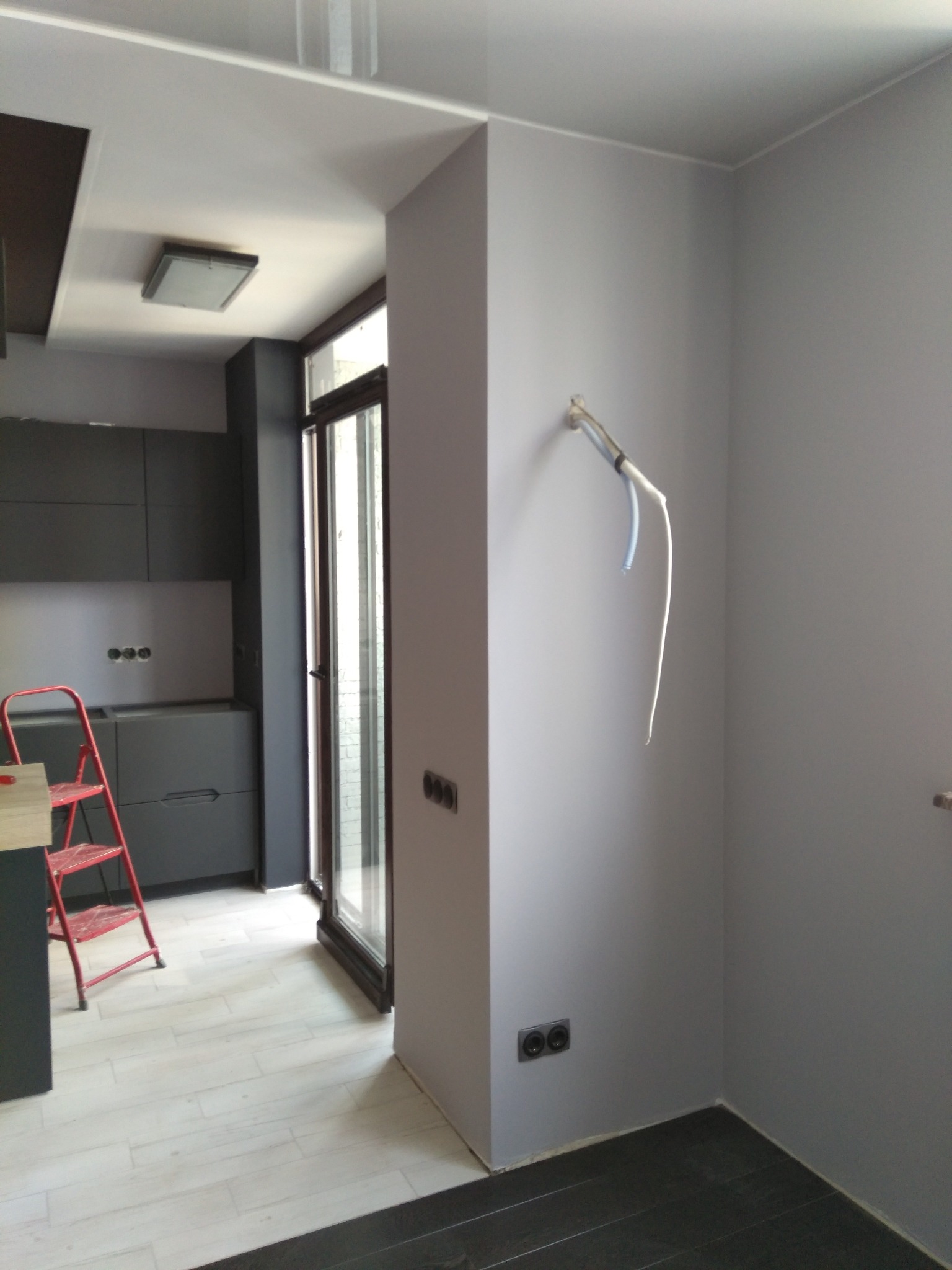 Szara kuchnia po remoncie: ściany pomalowane na szaro, widoczne gniazdka, fragment instalacji, drabina malarska, okno balkonowe i podłoga z paneli.