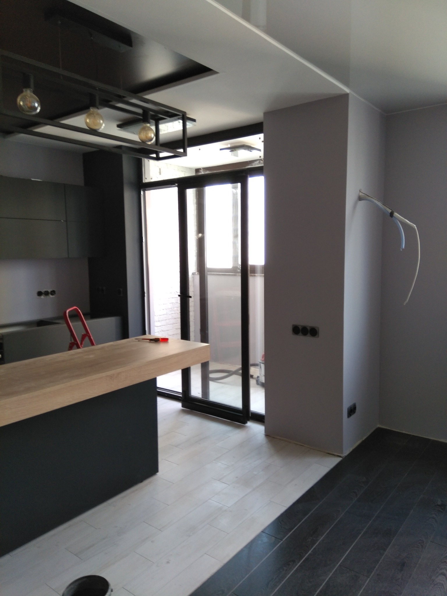 Nowoczesna kuchnia w trakcie wykańczania: szare ściany, drewniany blat, czarne meble i lampy. Widoczne przewody elektryczne i drabina.