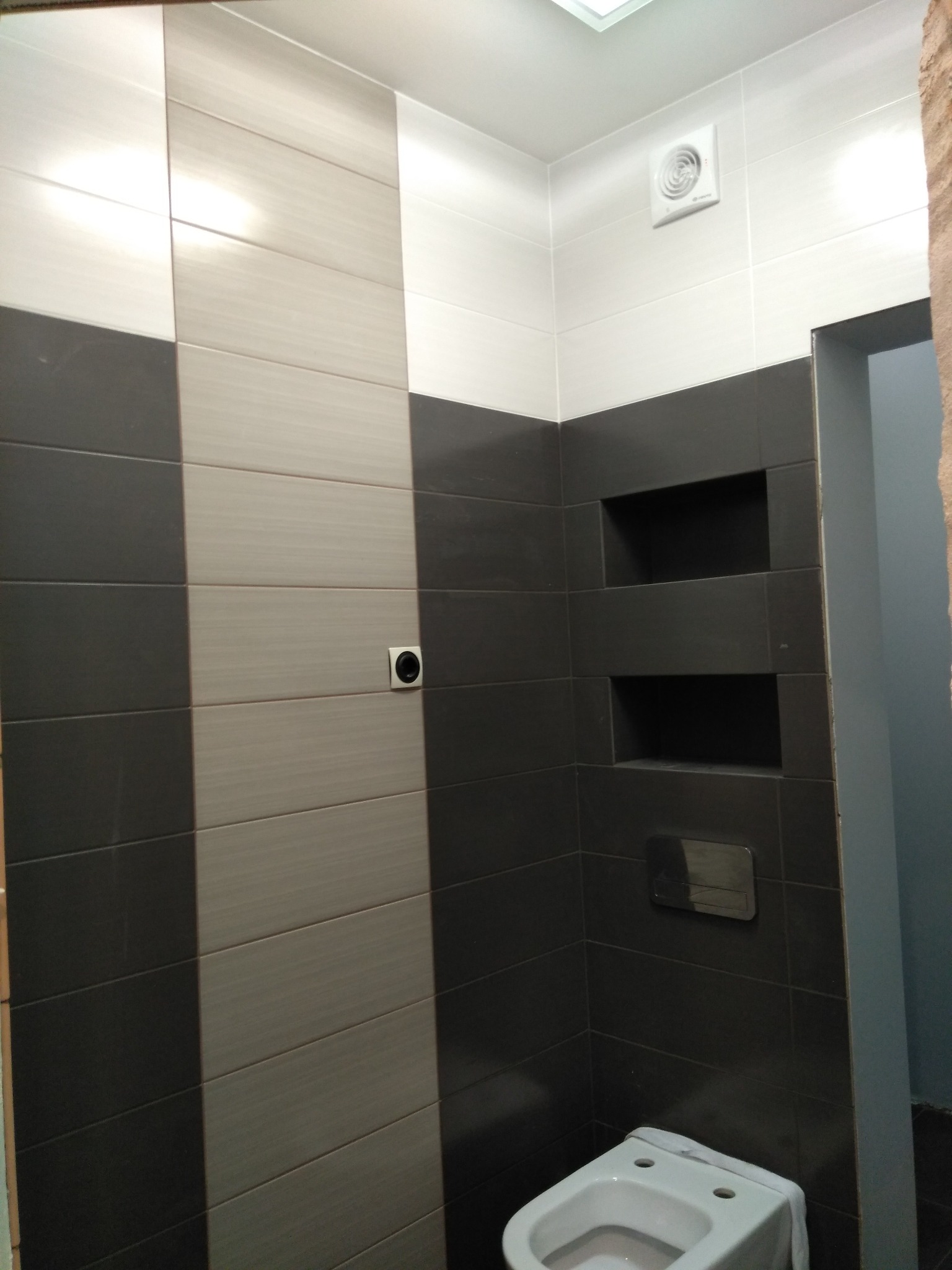 Wykończona łazienka z szaro-białymi płytkami ułożonymi w poziome pasy, wnęki w ścianie i biała toaleta. Widoczny fragment drzwi.