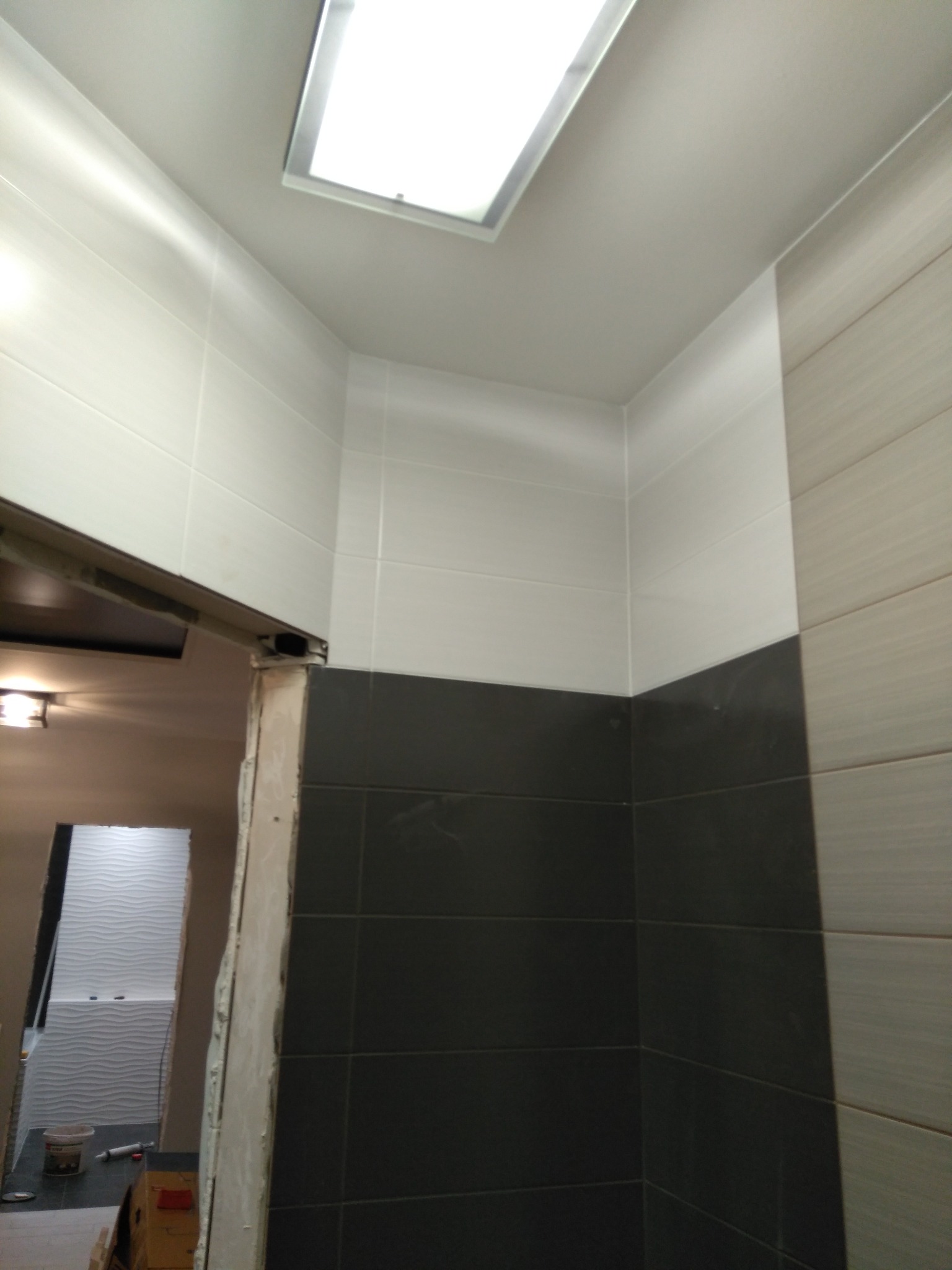 Wykończona łazienka z dwukolorowymi płytkami (białe i grafitowe) ułożonymi do połowy ściany. Widoczny fragment sufitu z oświetleniem i fragment drugiego pomieszczenia.