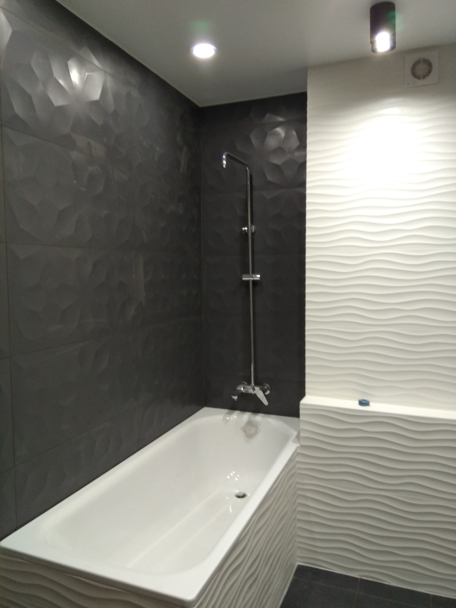 Nowoczesna łazienka z wanną obłożoną falistą płytką, ściany wykończone strukturalną, ciemną płytką. Chromowana bateria natryskowa, oświetlenie punktowe.