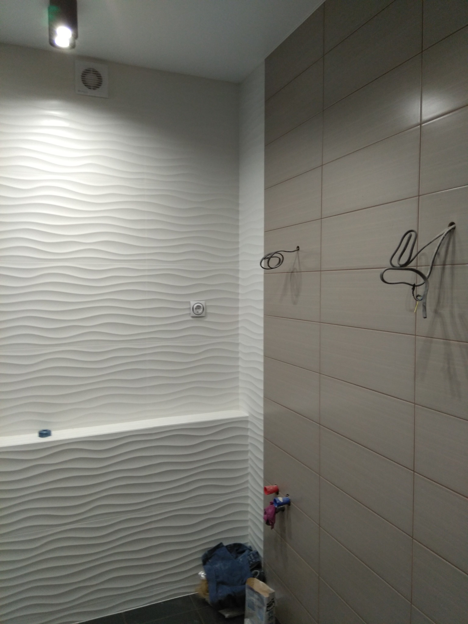 Wykończona łazienka z falistą, białą ścianą i beżowymi płytkami. Widoczne wystające kable i rury, sugerujące trwające prace wykończeniowe. Nowoczesny design.