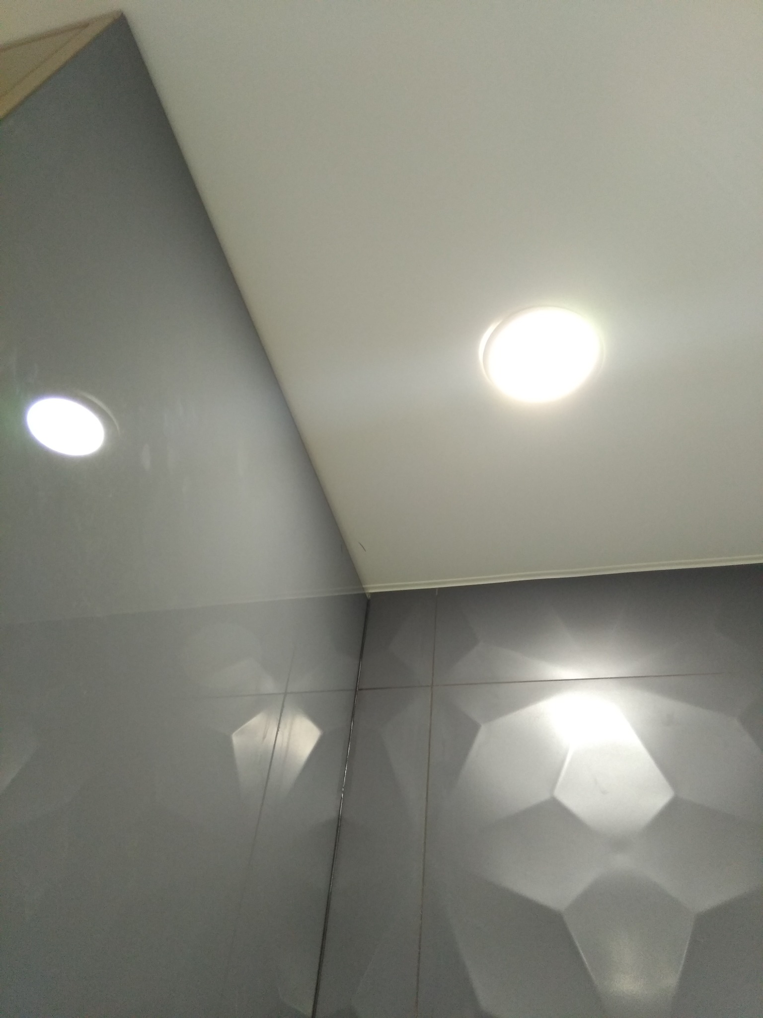 Nowoczesna łazienka z szarymi, trójwymiarowymi płytkami ściennymi i punktowym oświetleniem sufitowym. Minimalistyczny design, geometryczne wzory, jasne światło.