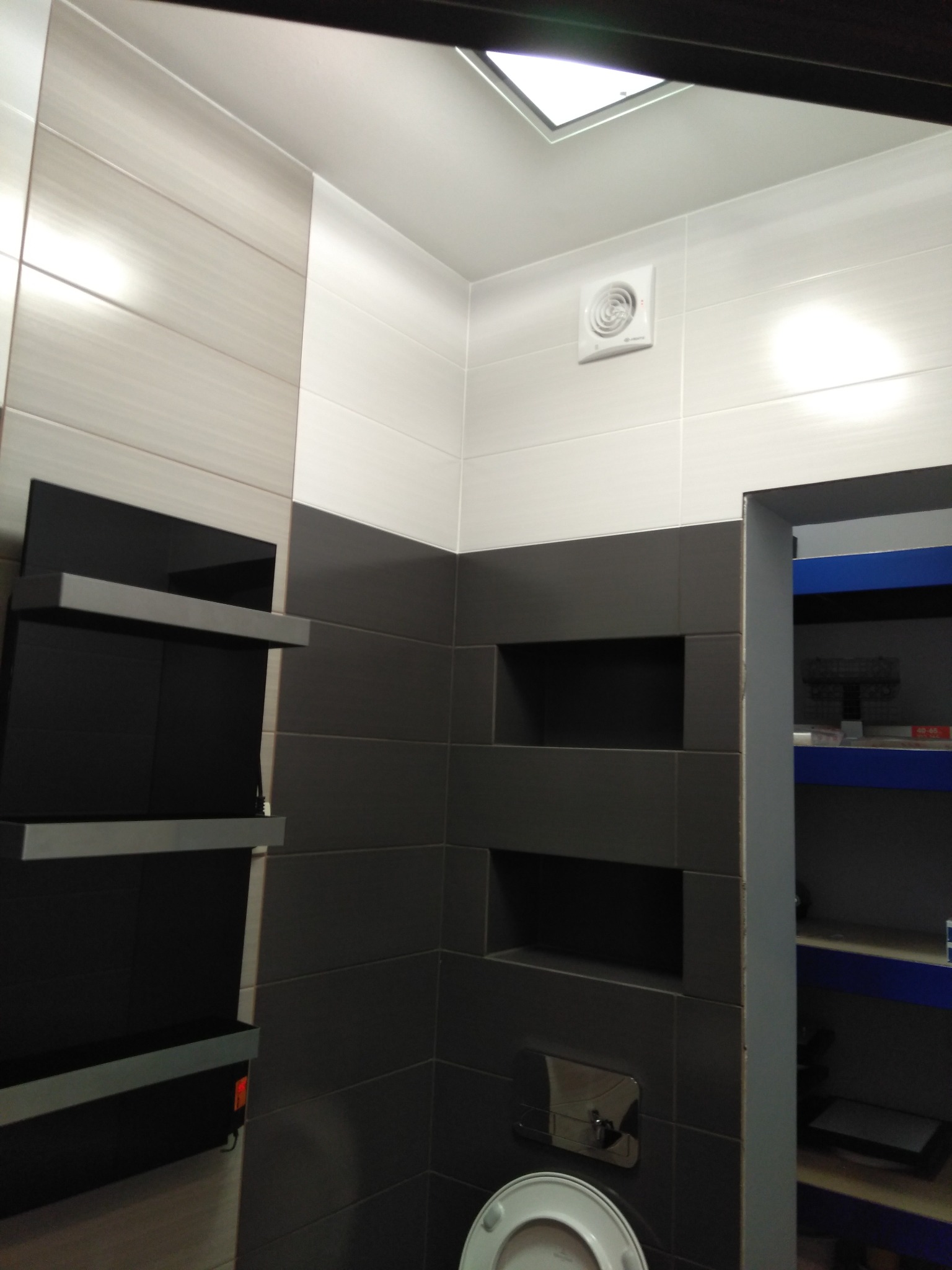 Nowoczesna łazienka z szarymi i białymi płytkami, wnękami w ścianie i podwieszaną toaletą. Widoczny wentylator sufitowy i półki.