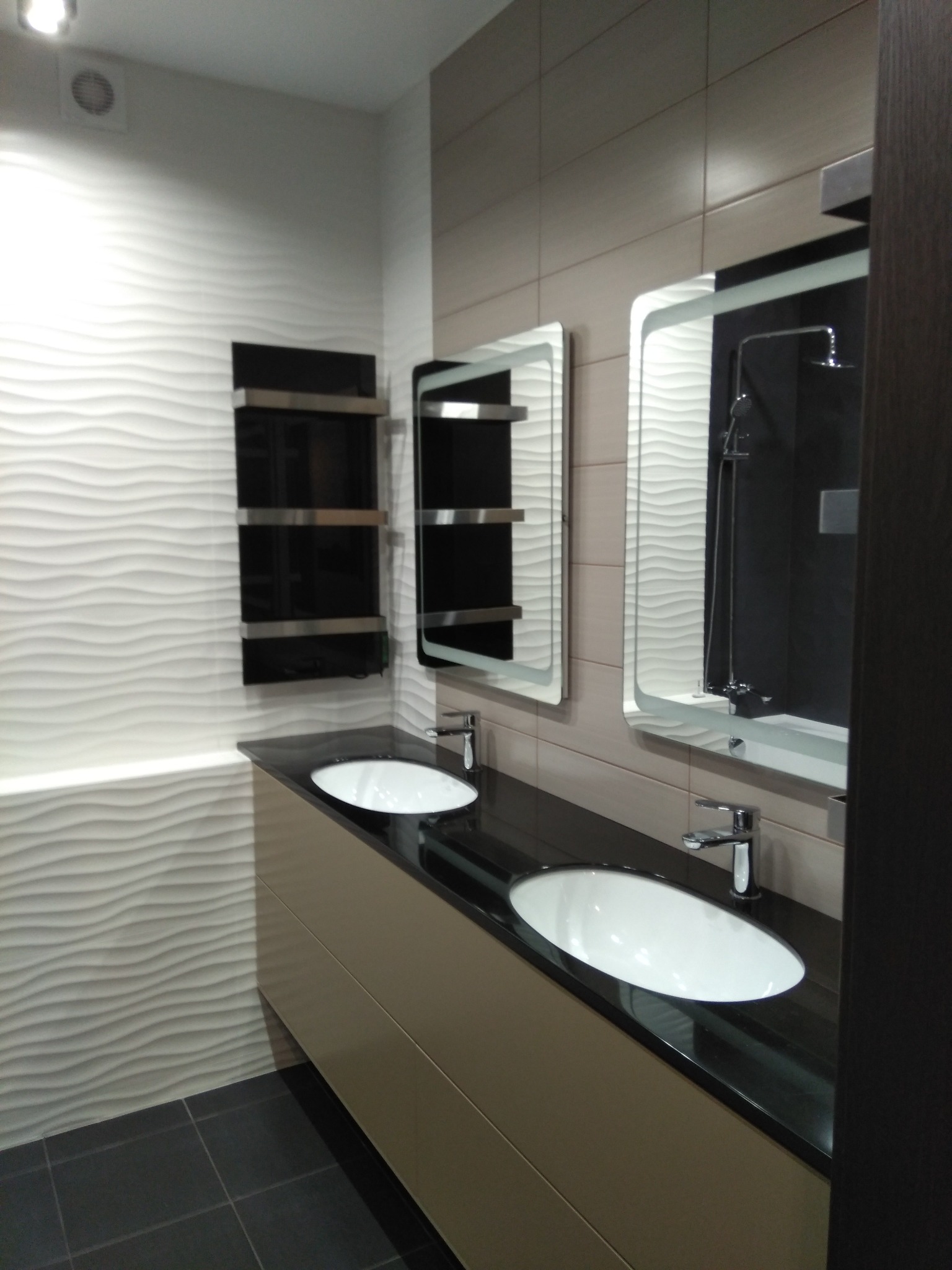 Nowoczesna łazienka z dwoma umywalkami na blacie z czarnego kamienia, podświetlane lustra i grzejnik drabinkowy na ścianie z dekoracyjną fakturą.