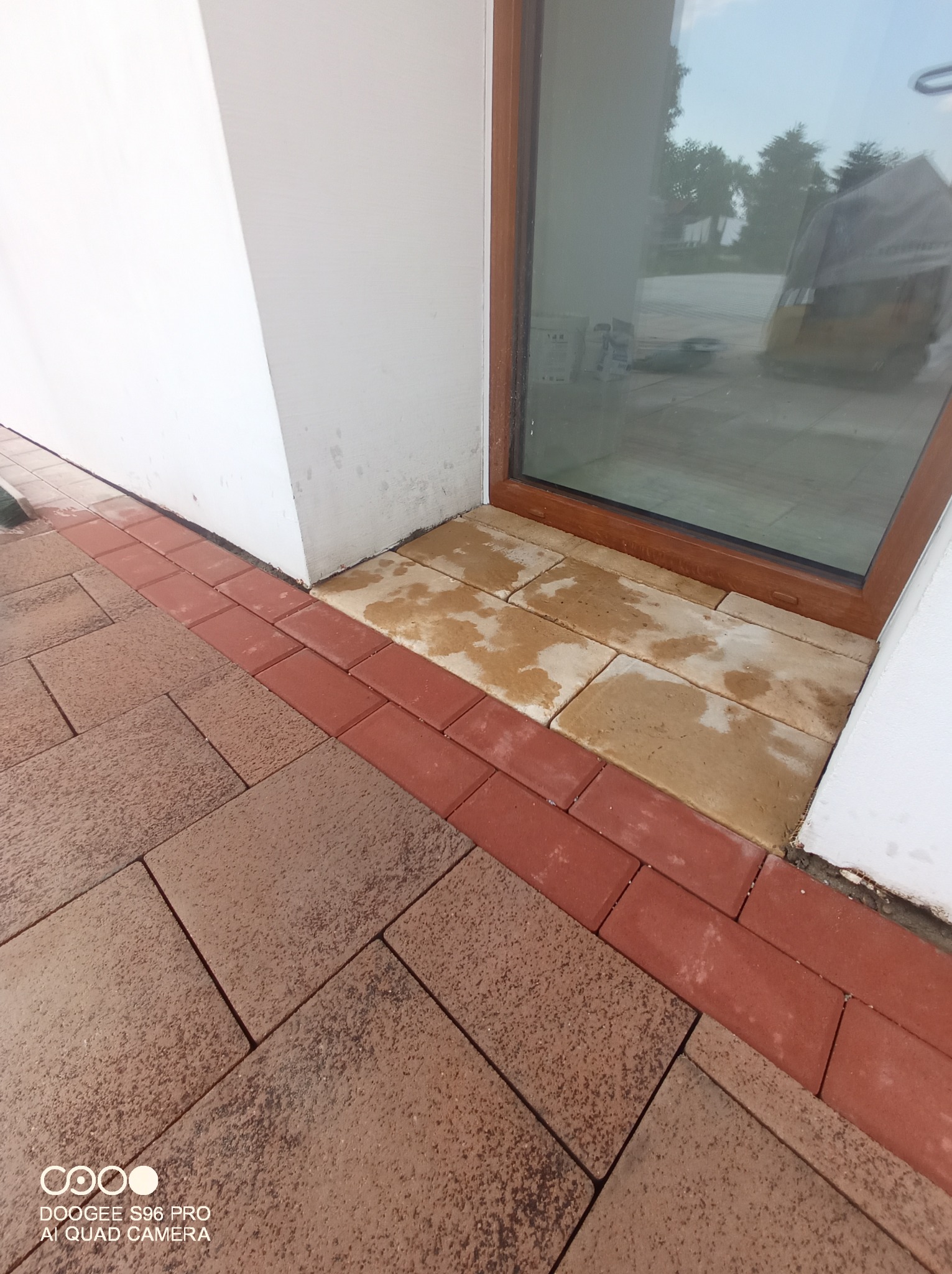 Ułożona kostka brukowa w kolorze brązowym i czerwonym obramowaniu przy oknie budynku, widoczne ślady wilgoci na kostce przy oknie.