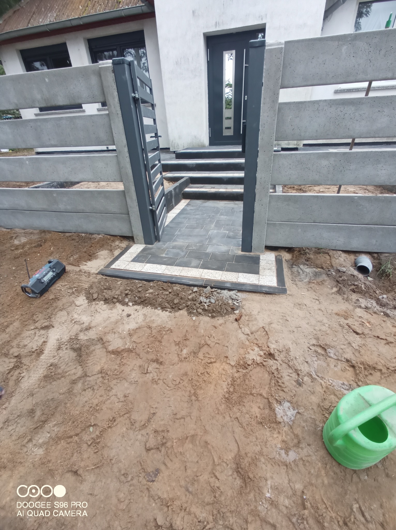 Nowoczesne, betonowe ogrodzenie panelowe z metalową furtką i ścieżką z szarej kostki brukowej z jasną obwódką, prowadzącą do wejścia do domu. Na pierwszym planie widoczna ziemia i narzędzia.