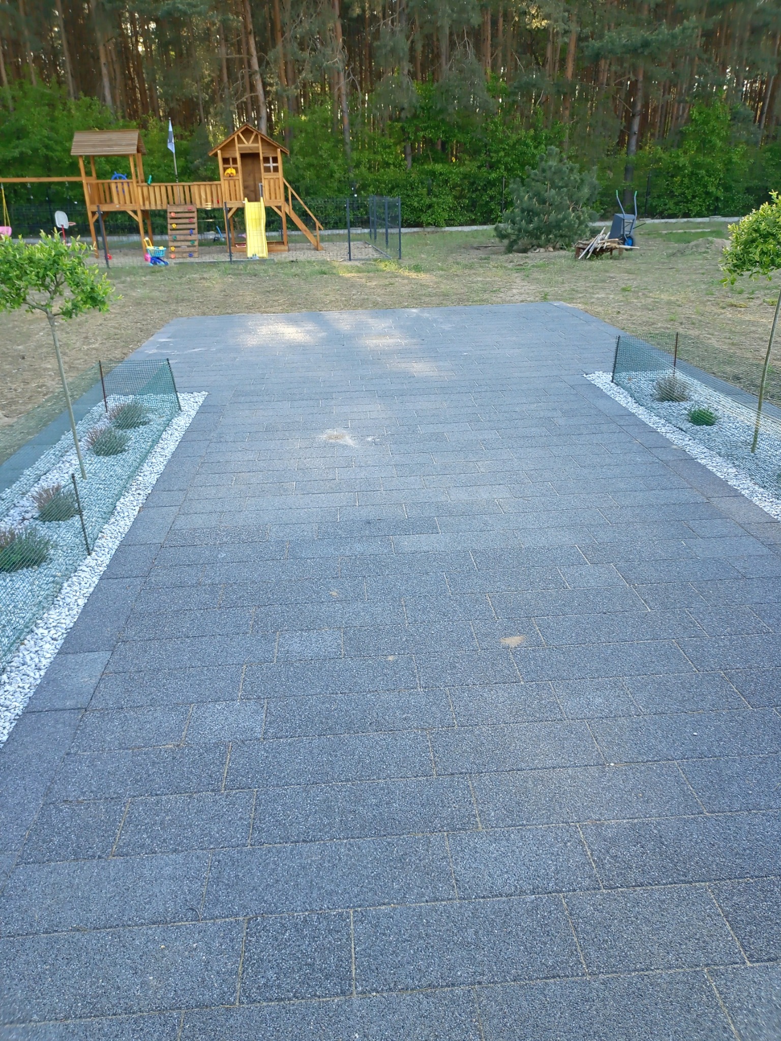 Szary, prostokątny plac z kostki brukowej otoczony białym żwirem i niską siatką ogrodzeniową, widok na plac zabaw i las w tle.